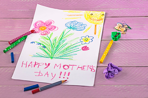 Children's drawing of Mother's Day greetings, pencils and paper on a pink wooden background. Happy Mother's Day concept.