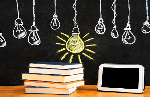 Old books, tablet on desk and blackboard. The chalkboard drawing shows light bulbs in a row with a big one glowing. The big light bulb is a symbol of success or intelligence. Copy Space.