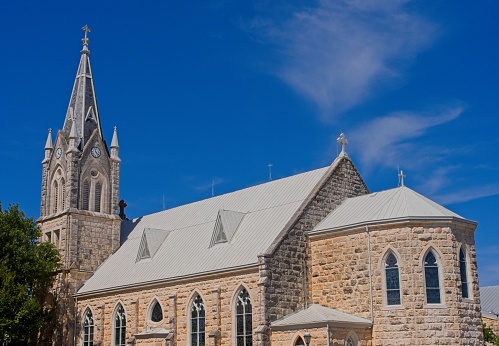 horizontal image of a quaint little white country church with a steeple sitting in a green meadow surrounded by trees under a beautiful blue sky with white clouds floating by in the summer time.