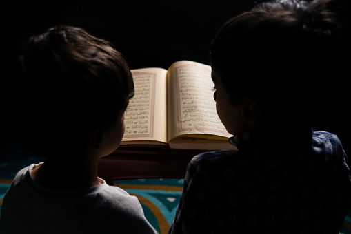 Younger brothers reading Quran sitting in mosque. The younger brothers are sitting on the carpet under the light coming from the mosque window. They are reading the Quran on the podium. They worship. Shot with a full frame camera.