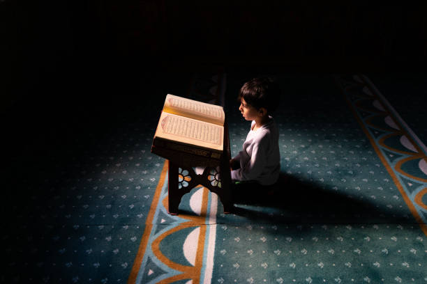 Boy sitting in mosque reading Quran. The child is sitting on the carpet under the light shining from the window of the mosque. He is reading the Quran in the lectern. worshiping. Shot with a full frame camera.