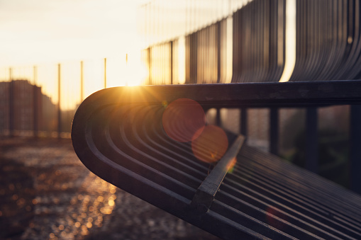 In the late evening of winter, a metal park bench stands alone on a cobblestone ground. The afterglow of the sunset spills over the bench bringing a sense of loneliness and silence.