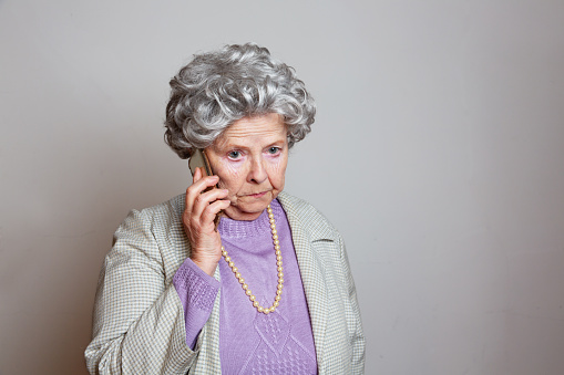 Eccentric senior woman,wearing oldfashioned purple pullover and pearl necklace, on the phone serious expression, could be just gossip