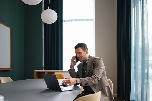 Handsome smiling man in a suit, sitting at the office, talking on his mobile phone while typing on the keyboard of his laptop computer.