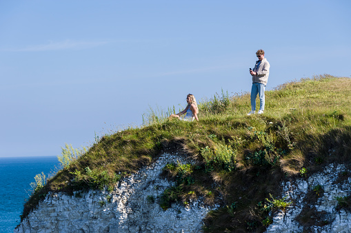 Studland, United Kingdom: 15th June 2021: A man takes a photo using his smartphone of his girlfriend who sits near the cliff edge on a hot sunny summer day in late afternoon in  Dorset.