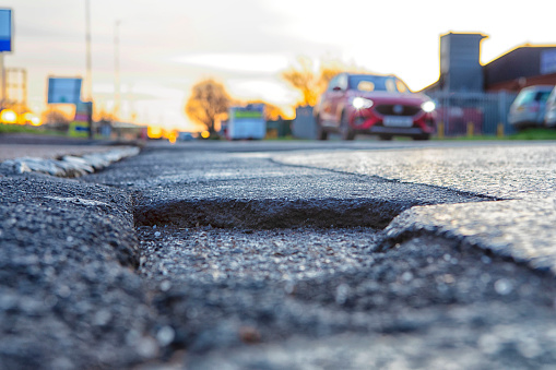 Potholes in the road with an approaching car (blurred, no identity). Potholes are a political hot potato in many countries as vehicles get heavier (electric batteries), traffic increases, and infrastructure maintenance budgets are squeezed, potholes are getting worse.