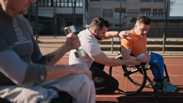 SLO MO Rack Focus of Fitness Coach Assisting Adaptive Athletes on Wheelchairs Lifting Dumbbells on Sports Ground