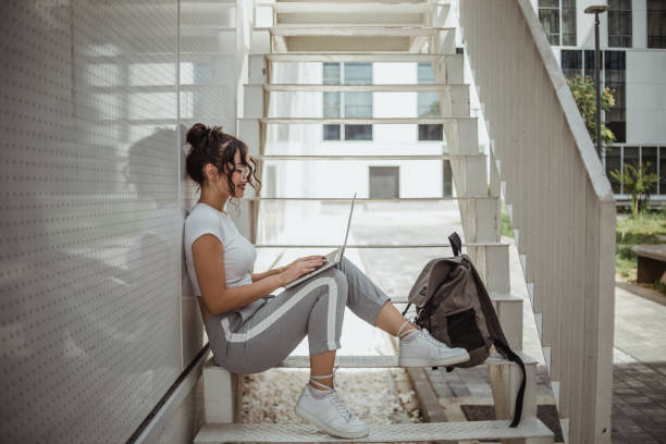 Portrait of a female student in casual clothes, sitting in front of the faculty building and using a laptop