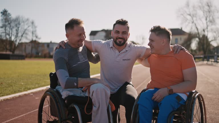 SLO MO Portrait of Smiling Fitness Coach with Arm Around Adaptive Athletes on Wheelchairs Looking At Camera on Sports Ground