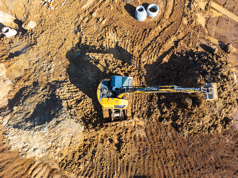 Aerial view of an excavator on a construction site with visible tracks in the dirt and construction materials nearby.