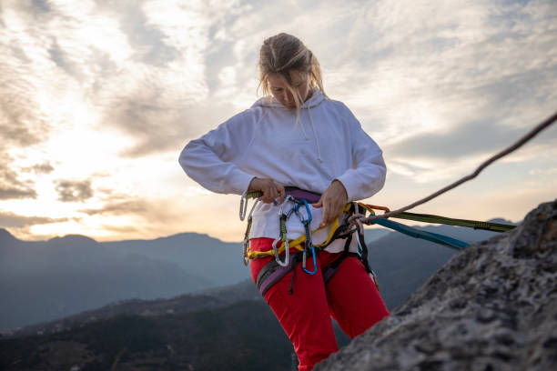 Young woman wearing in safety harness with climber equipment and helmet holding rope and looking at camera on background of rock
