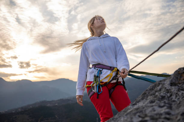 Young woman climber leans on rope and looks up on mountain cliff at sunset