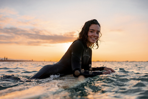 Attractive Middle Eastern female surfer resting on a surfboard in the middle of the ocean and waiting for a wave. The sun is setting and the sky is orange. The woman is happy and enjoying her summertime activities in Barcelona. She is smiling at the camera.