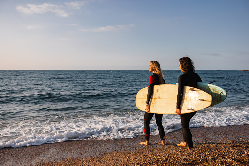 Two female surfer friends helping each other carry their surfboards. They are walking on a sandy beach in Barcelona on A beautiful summer morning after catching some waves. They are wearing black wet suits. The sun is illuminating the surfboard and their faces.