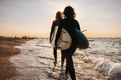Back view of two female surfers in wet suits walking one behind another and helping each other carry the heavy surfboards. They are returning from a fun afternoon surfing session. They have messy hair from the wind.