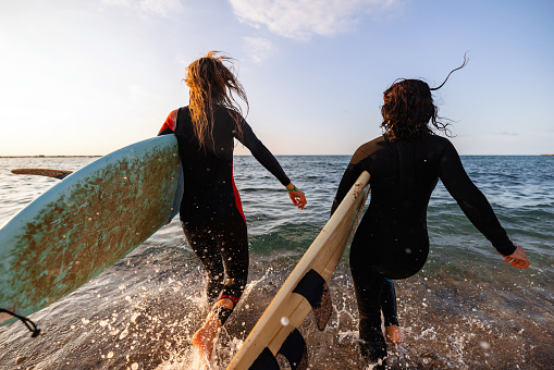 Two female friends going on a surf session together. They are running into the water while carrying their surfboards. The surfers are wearing black wetsuits. Fun summer adventures with friends.