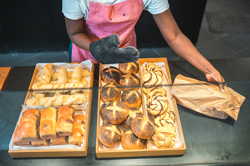 Close up on hand of a Black female shop assistant in an artisan bakery. Standing at a counter preparing order. Putting freshly baked goods in paper bags.