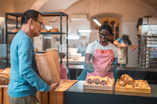 Mature Asian male selecting fresh bread from a modern bakery. Black female baker is taking her order over the counter.