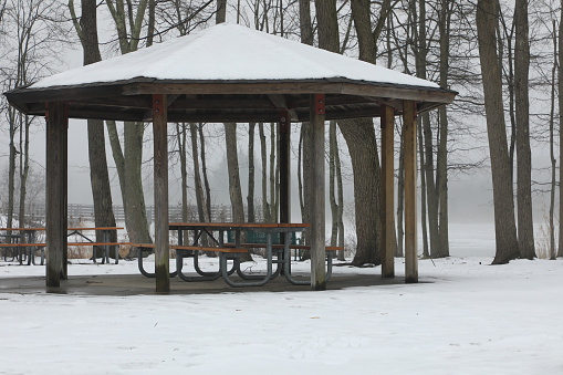 A park Gazebo in the snow. Snow on the ground and on top of the roof. Sitting along side a frozen lake on a misty foggy day.