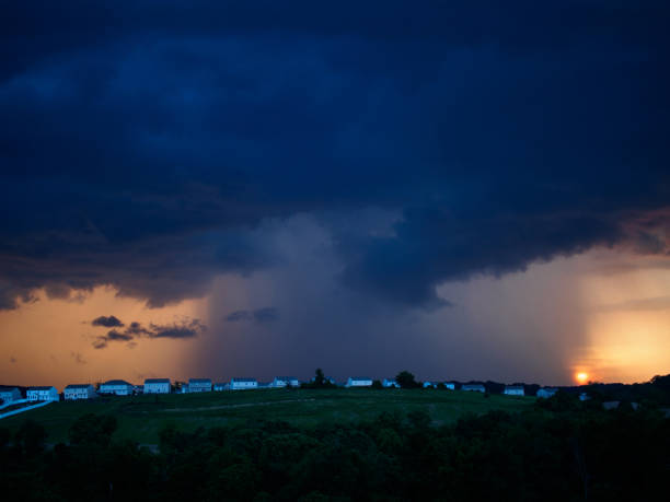 istock Dark clouds and turbulent sky with lightning and storm approaching and light at sunset over suburb houses on a green hilltop. 1983838091