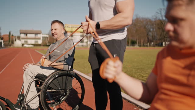 SLO MO Personal Trainer Assisting Paraplegic Athletes on Wheelchairs Doing Exercise with Resistance Bands on Race Track