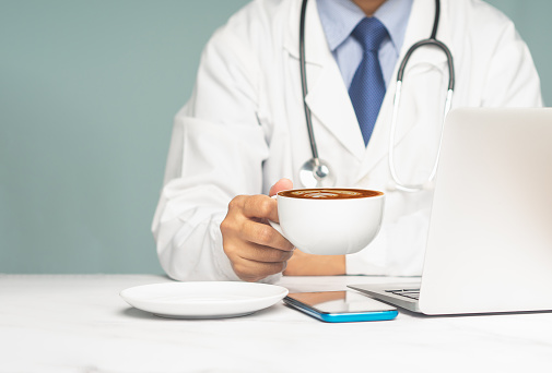 Doctor in uniform with a stethoscope holding a coffee cup while sitting at the desk. Medical and relaxation concept. Midsection. Close-up photo