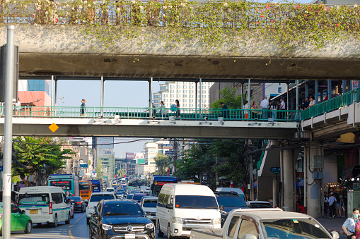 Thai people are walking in footbridge and traffic below BTS Skytrain at Vicotory Monument in Bangkok. Footbridge is over street Thanon Phaya Thai, Ratchathewi, Bangkokand is leading towards a mall and towards skytrain station.. On street is traffic jam entering into roundabout and square Victory Monument