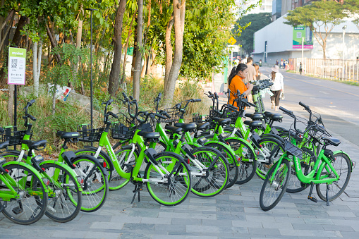Rentable city nicycles at corner of Chulalongkorn University Centenary Park in Bangkok. Bicyc kles are green colored. A person is standing at and behind bicycles and is using mobile phone. Behind woman are ev scooters. In background some people are walking,. Scene is at Banthat Thong Road, Wang Mai, Pathum Wan