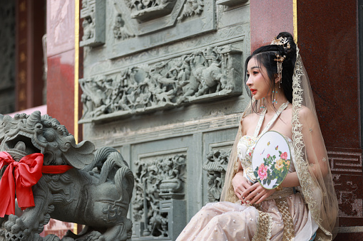 beautiful Asian young woman wearing  traditional Chinese cheongsam decoration sitting near the lion statue  at the shrine in Thailand for the Chinese New Year Festival