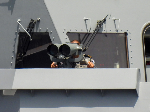 A sailor on watch looks through the binoculars on the bridge of HMAS Sydney, a Hobart Class destroyer of the Royal Australian Navy docked at Garden Island naval base in Sydney Harbour. His name badge has been blacked out to protect their privacy. This image was taken on a hot and sunny afternoon on 3 February 2024.