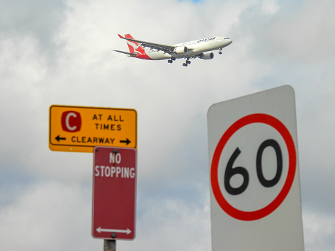 A Qantas Airbus A330-203 plane, registration VH-EBJ, coming into land on the third runway of Sydney Kingsford-Smith Airport as flight QF541 from Brisbane. In the foreground are road signs on Airport Drive, Mascot. This image was taken from near Nigel Love Bridge, Mascot on a windy and cloudy afternoon on 2 February 2024.