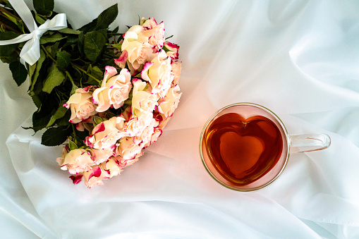 Heart-shaped glass teacup filled with red tea on white satin fabric, a bouquet of light pink roses at top left of picture,  horizontal