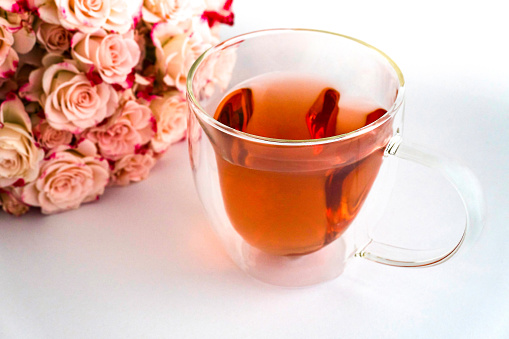 Heart-shaped glass teacup filled with red tea on white satin fabric, light pink roses on the left and a white sticky note reading Good morning Happy Birthday I love you pictured, top view, horizontal,  a bouquet of light pink roses at top left of picture,