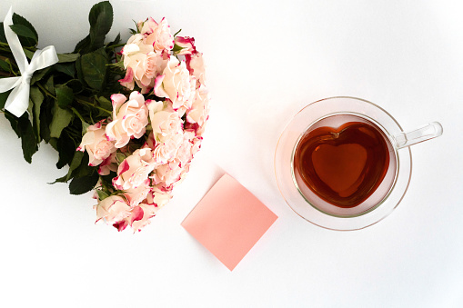 Heart-shaped glass teacup filled with red tea on a white background, a bouquet of light pink roses on the left and a blank pink note in the picture, top view, horizontal