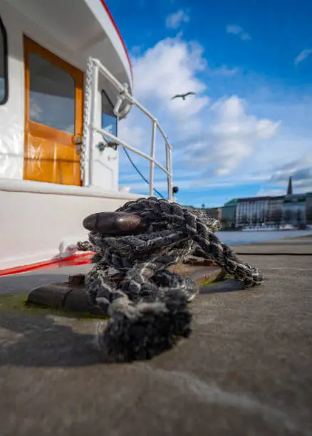 Cleat to which a black rope is attached, part of a white and red ship can be seen behind and buildings on the Alster and blue sky can be seen in the background, vertical