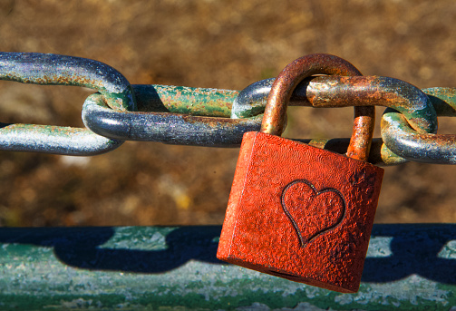 Single red padlock with heart on it connected to a chain, brown blurred background, horizontal