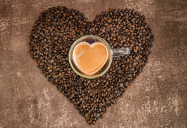 Heart-shaped glass cup filled with coffee on a heart of roasted coffee beans Heart-shaped glass cup filled with coffee on a heart of roasted coffee beans against a mottled light brown background, horizontal, top view tasse café stock pictures, royalty-free photos & images