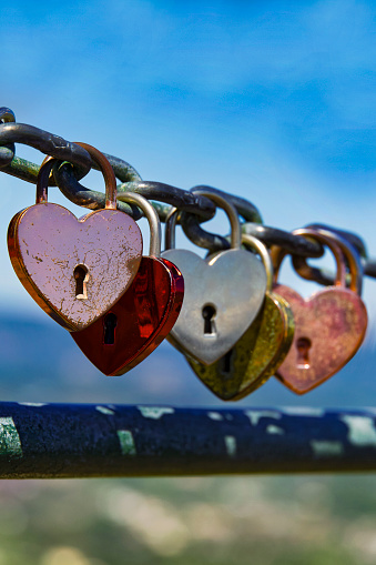 Five different colored heart shaped padlocks hanging from a chain in a row, light blue sky background, vertical