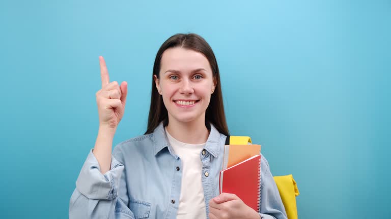 Portrait of pretty young woman student in shirt with backpack holding books point index finger overhead workspace area mock up, posing isolated on blue background. Education in high school concept