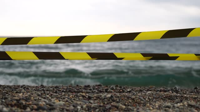 Yellow black warning tape barrier ribbon swinging in the wind across exotic sea beach background without people. No entry Yellow black caution tape. No holiday concept, delayed travel, no summer plans