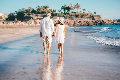 Romantic senior couple walking barefoot on the seashore at sunset enjoying vacation and retirement, two smiling people expressing love and tenderness