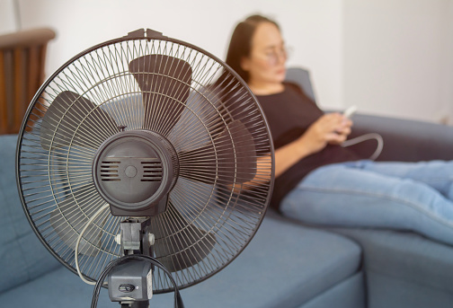 Close up shot of electric fan on floor at home. Closeup of black electric fan heater on floor in living room, with woman on couch in background. Climate, temperature in the house concept
