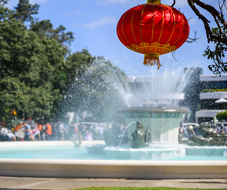 Chinese New Year lantern hanging under the Pohutukawa tree. Fountain and unrecognizable people in the background. Mission bay. Auckland.