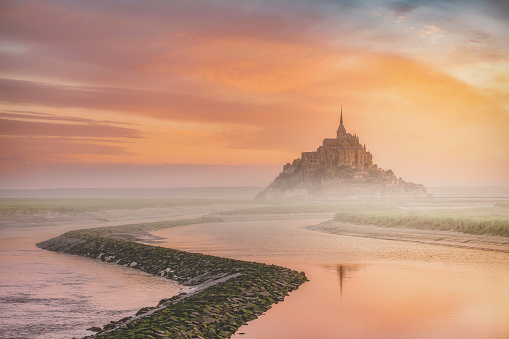 Beautiful view of the famous Le Mont Saint Michel. East across the tidal bay from the salt marsh at a beautiful early morning colorful cloudy sunrise time in Normandy, Northern France