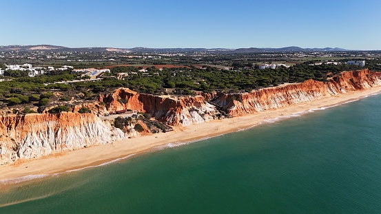 Aerial view of the south of Portugal in wonderful region called Algarve, daytime on a sunny clear sky day along the beach called Praia da Falesia with people relaxing and enjoying soft golden sand, clear waters, stunning coloured red and orange cliffs forested with pine trees