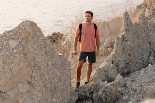 istock Handsome man in shorts with backpack staying on rocky coastline with warm sun light. 1984699684