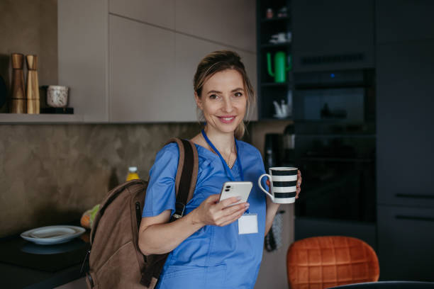 istock Female doctor getting ready for work, reading message on smartphone, drinking cup of coffee, leaving house in scrubs with backpack. Work-life balance for healthcare worker. 1984717355