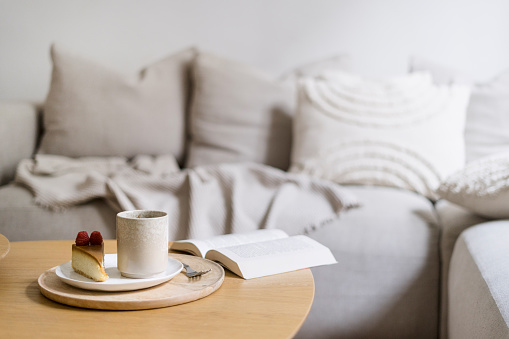 Selective focus on wooden tray with cup of coffee and sweet cake near open book on side table against comfort couch with pillows in cozy living room in morning. Breakfast concept. Relax at home