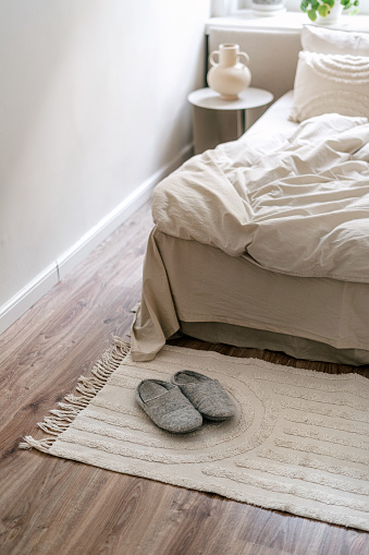 Selective focus on slippers standing on rug close to bed with bedding. Cozy bedroom with modern interior design. Concept of recreation in hotel, spending time at home