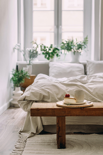 Still life details at cozy home. Close up of serving tray with breakfast on wooden bench against blurred background of bed and bright bedroom interior in bohemian chic style